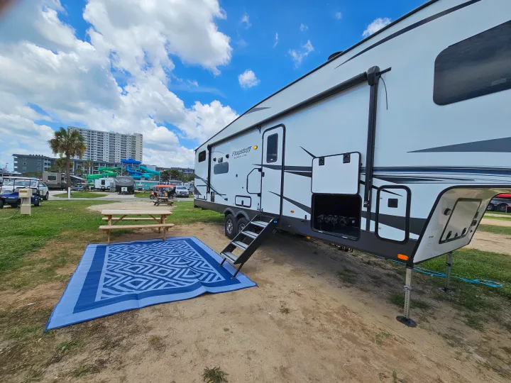 A modern Flagstaff RV rental is parked on a sandy campsite. A blue outdoor mat and picnic table are visible, with a water park in the background.. Forest River Flagstaff Super Lite 2022