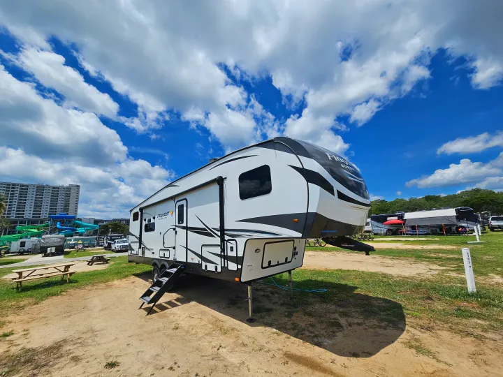 A modern white & black fifth-wheel RV rental is set up at a bustling campground with a water park, under bright blue skies.. Forest River Flagstaff Super Lite 2022