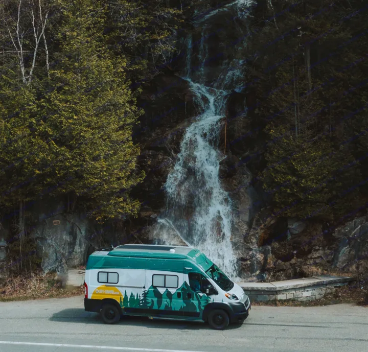An RV rental camper van with mountain graphics is parked by a road, with a majestic waterfall cascading behind it.. Winnebago Solis 2022