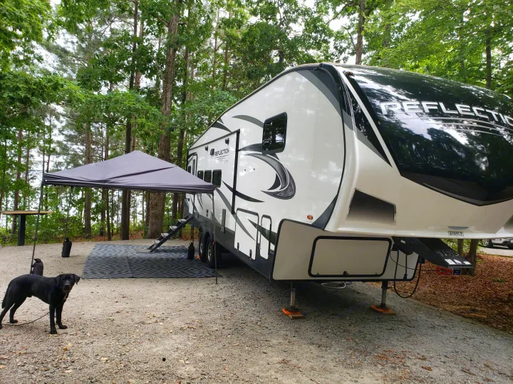 A large white and black Reflection fifth-wheel RV rental is parked on a gravel campsite under tall trees, with a black dog.. Grand Design Reflection 2021