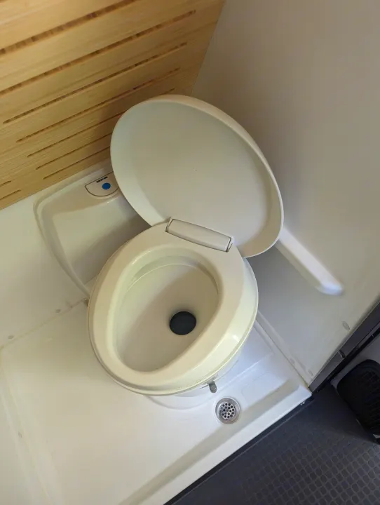 RV rental compact wet bath: Thetford toilet with open lid, shower drain, and wooden slatted wall details.. Winnebago Revel 2023
