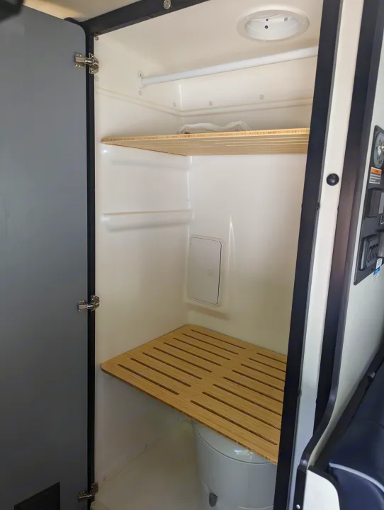 RV rental compact wet bath: toilet under slatted wooden bench, upper shelf, and shower rod. Practical interior for travel.. Winnebago Revel 2023