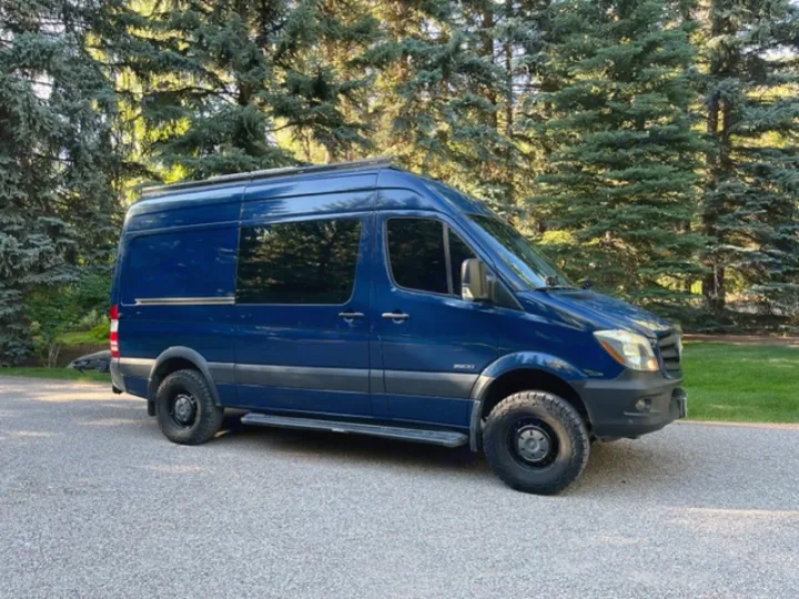 High-roof, dark blue Mercedes Sprinter camper van with rugged tires parked on gravel. An ideal RV rental for adventures.. Mercedes Sprinter 2016