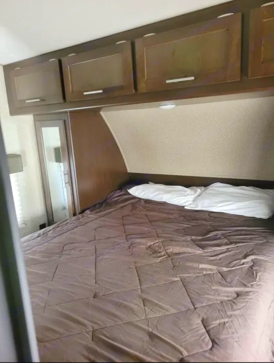 This RV rental bedroom features a bed with a brown comforter, white pillows, and dark wood overhead storage cabinets.. Forest River Wolf Pack 2016