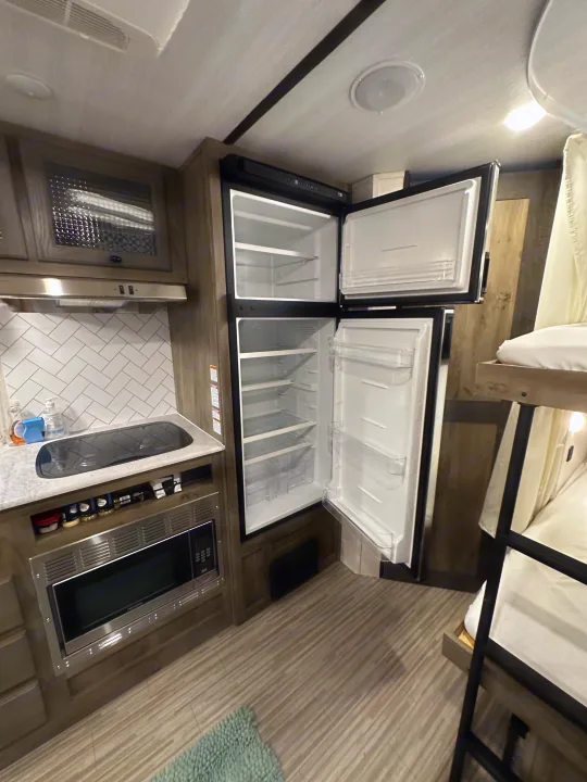 Inside an RV rental: a compact kitchen area with a cooktop & microwave, an open, empty refrigerator, and bunk beds.. Forest River Surveyor Legend 2021