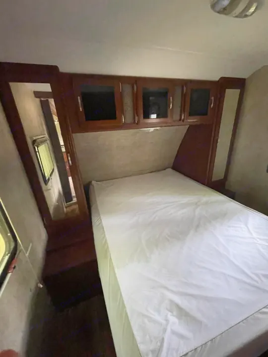 Inside this RV rental, a compact bedroom features a bed with white sheets, wood overhead cabinets, and a side window.. Keystone RV Passport Ultra Lite 2015