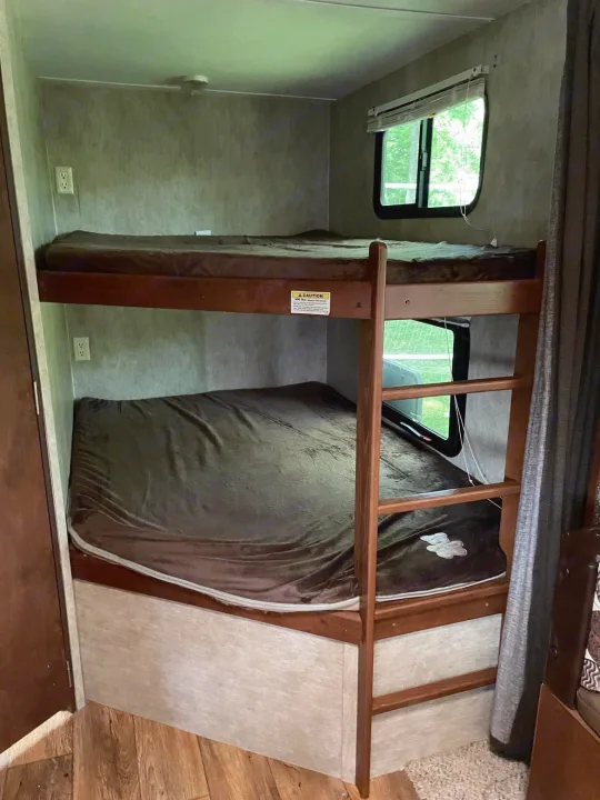 Inside an RV rental, two bunk beds with brown blankets offer a cozy, compact sleeping space, with a window and outlets visible.. Keystone RV Passport Ultra Lite 2015