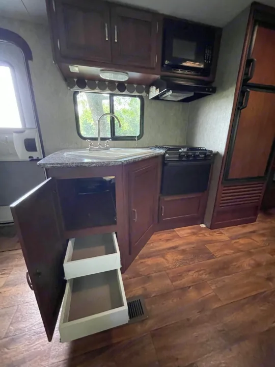 RV rental kitchen interior featuring dark wood cabinets, a sink, countertop, stove, microwave, and open storage drawers.. Keystone RV Passport Ultra Lite 2015