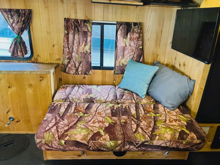 Rustic RV rental interior: wood-paneled walls, camo sofa/bed, multiple windows, and a wall-mounted TV for a cozy stay.. Forest River Cherokee Grey Wolf 2015