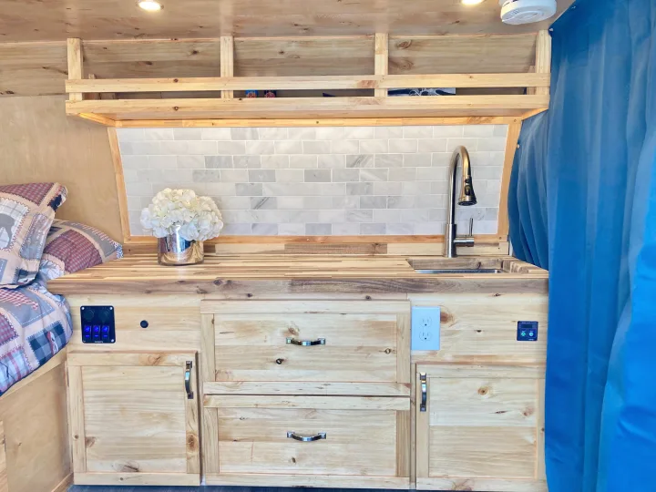 RV rental camper van interior: a well-equipped kitchen with wood counters, marble-look backsplash, sink, & built-in storage, next to a bed.. Ford Econoline 2012