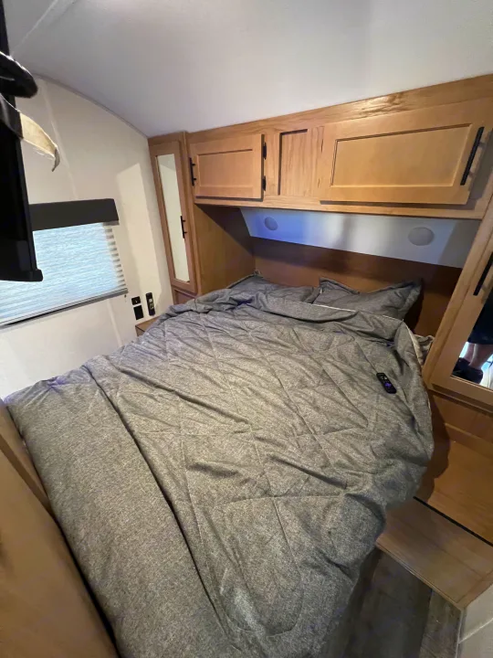 Inside an RV rental: a compact bedroom with a grey bed, overhead wood cabinets, a window, and small mirrors.. Forest River Cherokee Grey Wolf 26DBH 2023