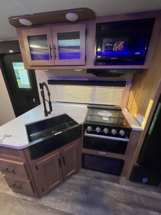This RV rental kitchen features a black farmhouse sink, a four-burner stove, a microwave, and illuminated upper cabinets.. Forest River Cherokee Grey Wolf 26DBH 2023