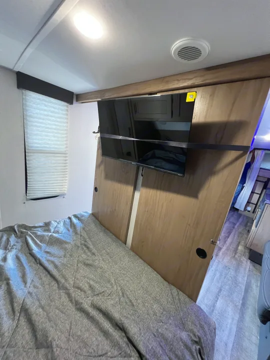 Inside an RV rental: a modern bedroom with a grey bed, wall-mounted TV, pleated window blinds, and overhead lighting.. Forest River Cherokee Grey Wolf 26DBH 2023
