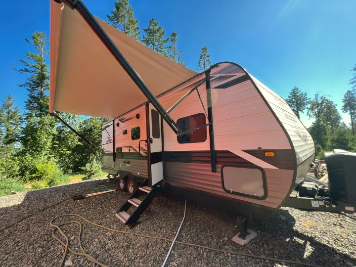 Sunny day showing a travel trailer RV rental with its awning extended, parked on gravel, surrounded by trees.. Jayco Jay Flight SLX Rocky Mountain Edition 2022