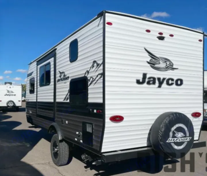 White Jayco travel trailer, seen from the rear-side, ideal for RV rental. Features black trim, mountain graphics, and a rear spare tire.. Jayco Jay Flight SLX 2023