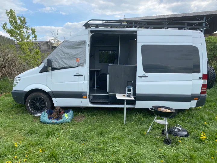 Enjoying an RV rental adventure: white van open to a grassy camp, with a dog, grill, and portable radio.. Mercedes-Benz Sprinter 2012