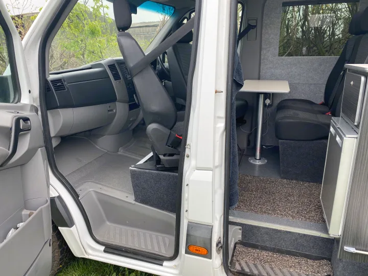 Experience this RV rental! Compact campervan interior showing a swiveling passenger seat, table, and integrated storage/fridge.. Mercedes-Benz Sprinter 2012