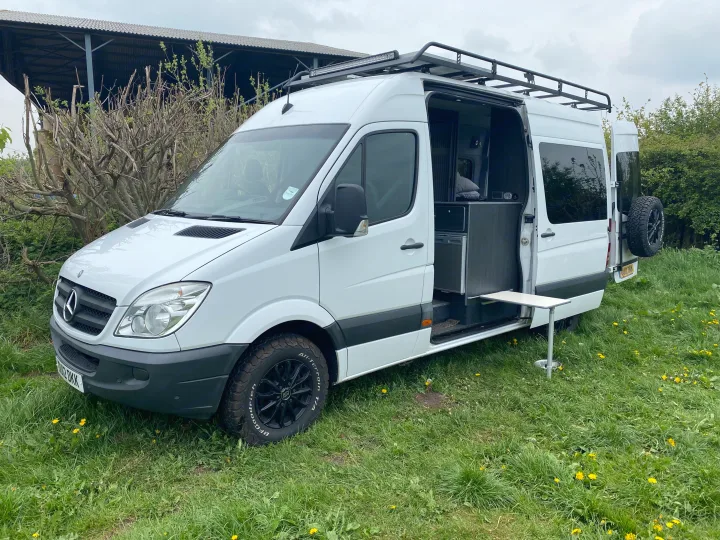 White Mercedes Sprinter **rv rental** campervan with open door, roof rack, all-terrain tires, parked in grass. Ready for adventure!. Mercedes-Benz Sprinter 2012