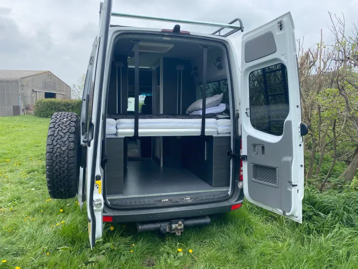 Rear view of an RV rental campervan showing its cozy bed setup, interior storage, and outdoor tire mount.. Mercedes-Benz Sprinter 2012