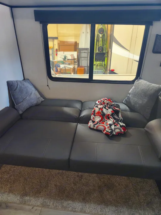 Inside an RV rental: a comfy sectional sofa with a colorful blanket. Window view of a stored kayak and RV in a facility.. Keystone RV Springdale 2022