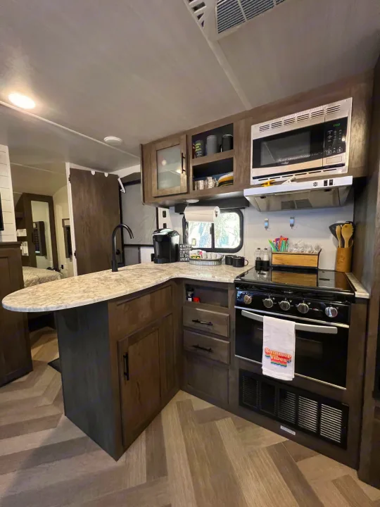 Modern RV rental kitchen with sleek island, stove, and microwave, offering a cozy bedroom view.. Forest River Wildwood X-Lite 2021