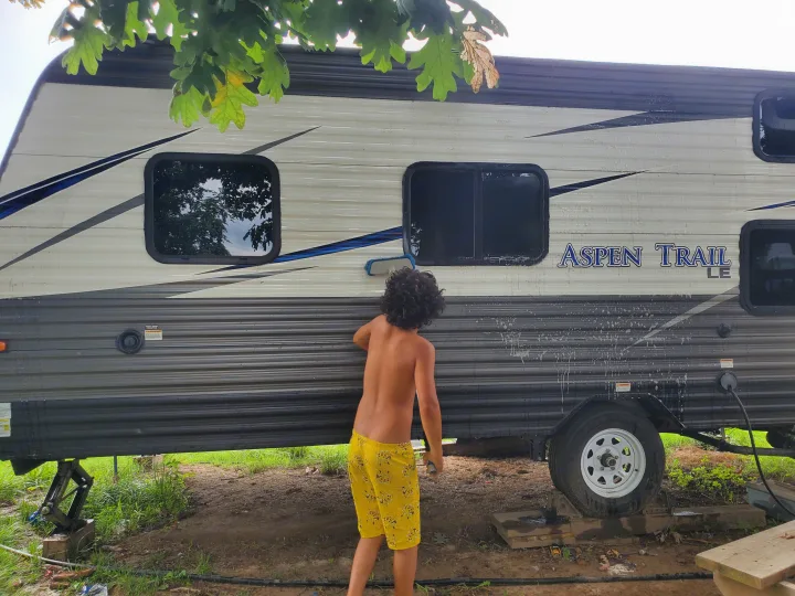 A young person with curly hair washes an Aspen Trail RV rental, scrubbing the camper's white and gray side with suds.. Aspen Trail Aspen Trail Trailer 2019