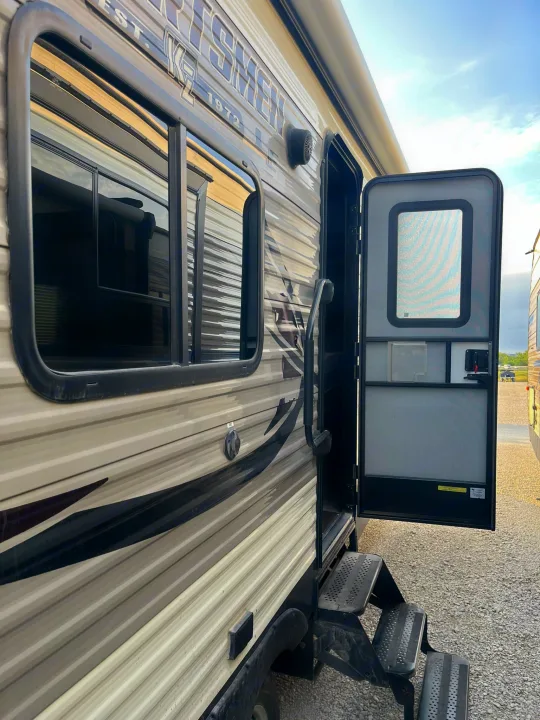 Side view of an RV rental: its door is open, steps extended, and windows reflect the surroundings.. Sportsmen LE 2018