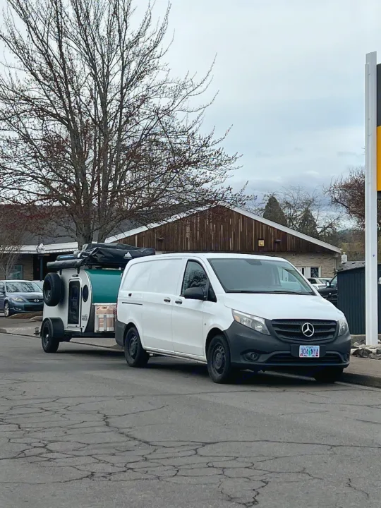 A white Mercedes van tows a compact, teardrop RV rental trailer on a street, ready for a road trip adventure.. Teardrops Northwest Trek 2022