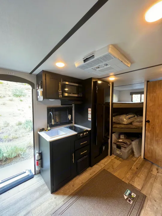 Bright RV rental interior showcasing a black kitchen with microwave, sink, stove, large fridge, and bunk beds. Ready for adventure!. Keystone RV Springdale 2023