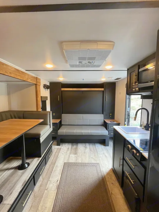 Modern RV rental interior with dining booth, sofa, and compact kitchen. Light wood floors and dark cabinets create a chic space.. Keystone RV Springdale 2023