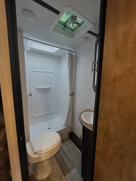Compact RV rental bathroom featuring a shower with curtain, toilet, sink, mirror, and skylight with fan.. Keystone RV Springdale 2023