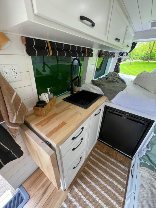 RV rental: Modern camper van interior shows a compact kitchen with wood counter, black sink/faucet, fridge, and bed.. Mercedes-Benz Sprinter 2017