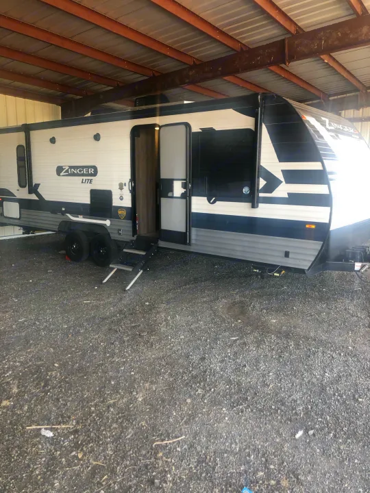 A white, black, and grey Zinger Lite travel trailer RV rental, with steps extended, sits under a metal shed on gravel.. Crossroads RV Zinger Lite 2022