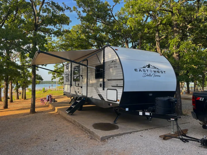 Relaxing by the lake in an rv rental. Modern travel trailer with an awning at a scenic campsite surrounded by trees.. East to West Della Terra 2022