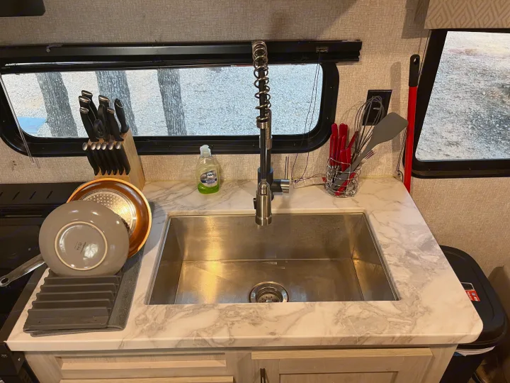 Modern RV rental kitchen with stainless sink, high-arc faucet, knife block, and organized utensils. View outside shows trees.. East to West Della Terra 2022