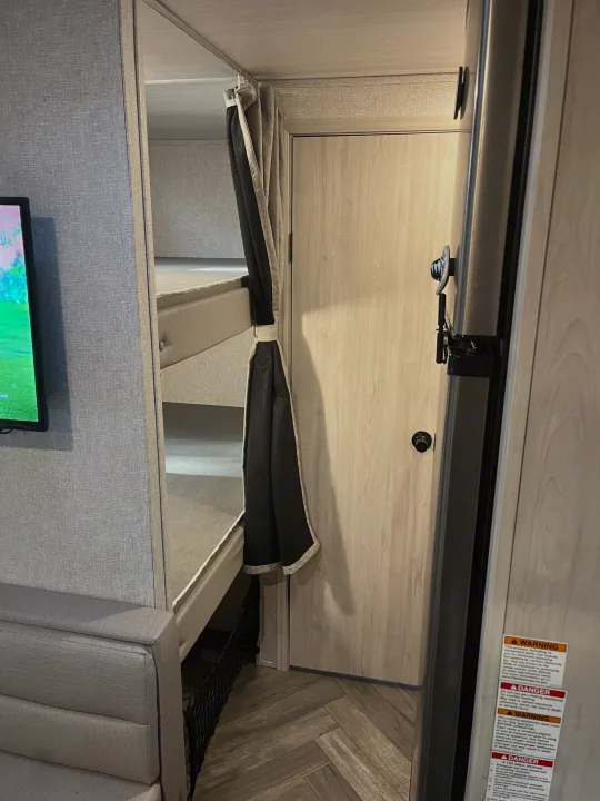 Inside an RV rental: cozy bunk beds with privacy curtain, TV, and light wood interior, perfect for compact living.. East to West Della Terra 2022