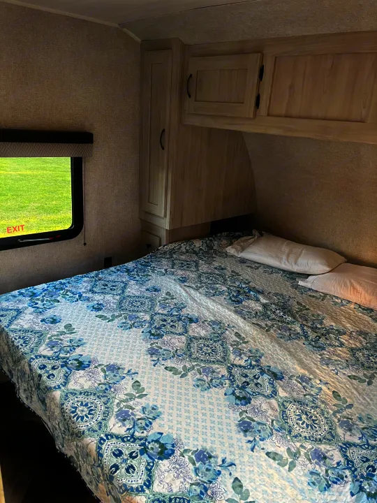 Cozy RV rental bedroom with floral bedding, wall cabinets, and a window overlooking a green field, perfect for travel.. East to West Della Terra 2022
