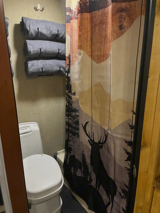 Cozy RV rental bathroom featuring a compact toilet, neatly rolled grey towels, and a scenic deer-themed shower curtain.. Forest River Cherokee Grey Wolf 2015