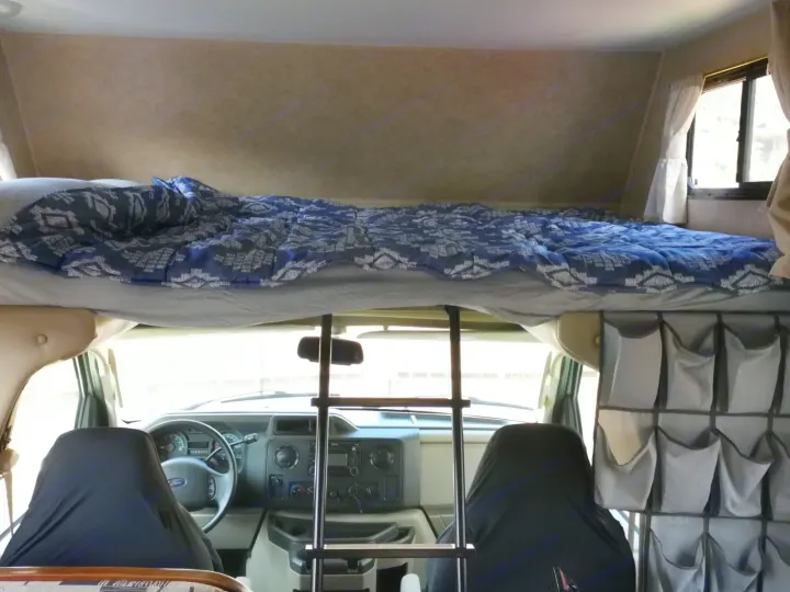 Cozy RV rental interior featuring an overhead bunk bed with bedding, a ladder, and front cab.. Forest River Sunseeker 2010