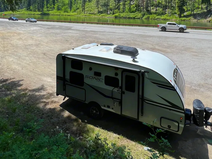 A white Sonic Lite RV rental is parked by a scenic road, lake, and forest, with cars driving by.. Sonic Lite 2022