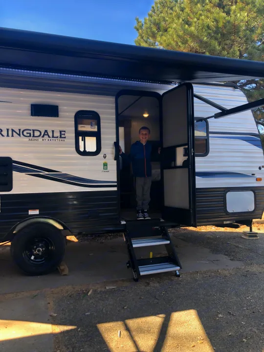 A smiling boy stands in the doorway of an RV rental, featuring its steps, awning, and outdoor setting.. Keystone RV Springdale 2022