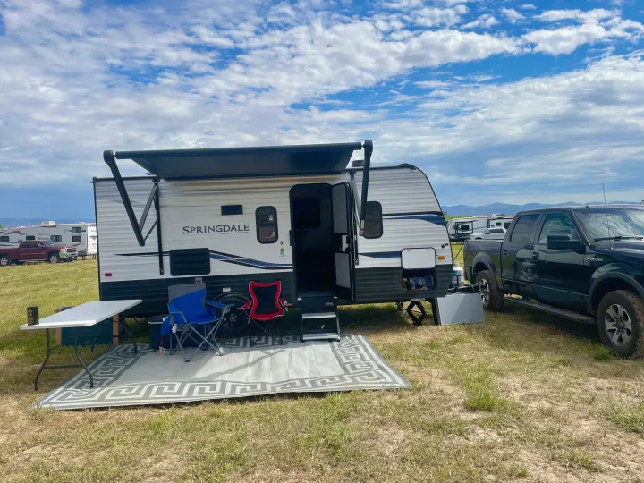 A white Springdale travel trailer with awning deployed, two chairs, and a black pickup truck at a grassy RV rental campsite.. Keystone RV Springdale 2022