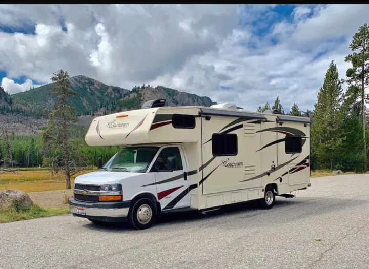 Cream & white Coachmen RV rental parked on a road. Mountains, pine trees & cloudy sky in this scenic outdoor setting.. Chevy 4500 2019