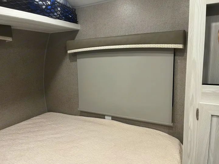 Inside an RV rental bedroom featuring a white bed, pull-down window shade, gray walls, and storage net.. Forest River Rockwood Geo Pro 2020