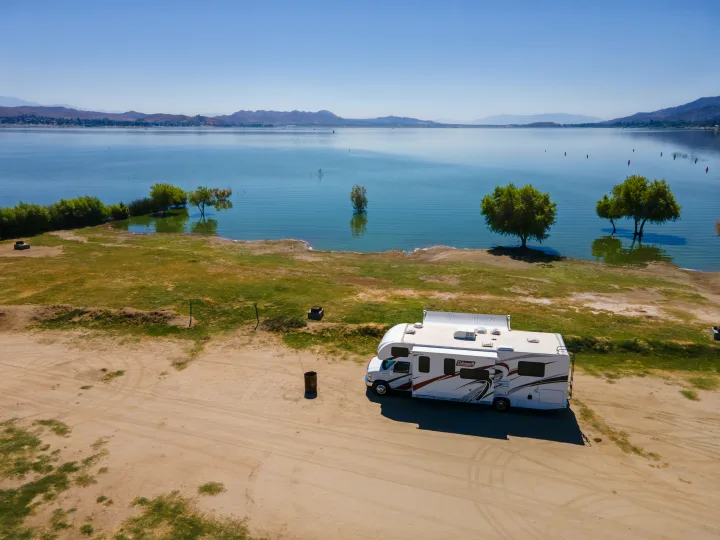 A white Coleman RV rental is parked on a dirt shore by a serene lake with submerged trees & distant mountains.. Thor Coleman 2023