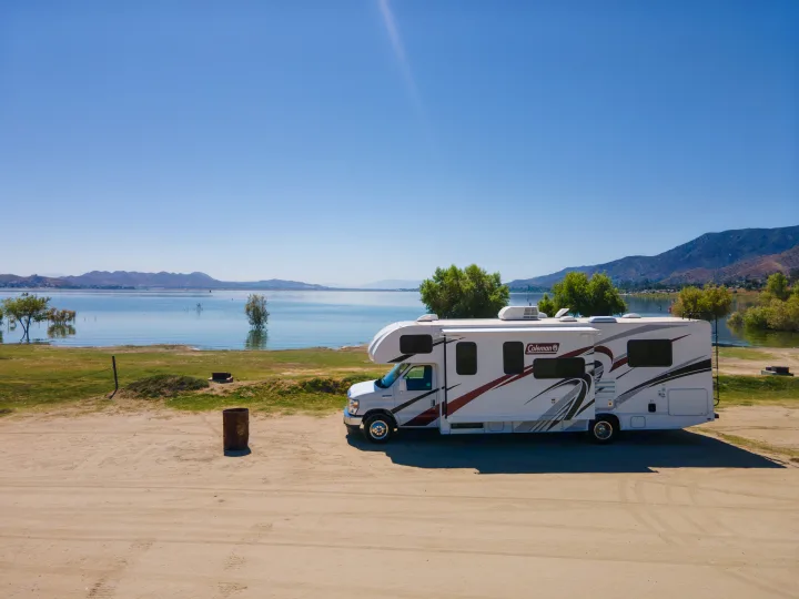 Scenic rv rental view: A white Coleman RV parked on a dirt lot beside a calm lake with mountains under a bright sky.. Thor Coleman 2023