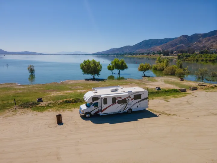 White RV rental parked at a serene lakeside campsite with trees and distant mountains under a clear blue sky.. Thor Coleman 2023