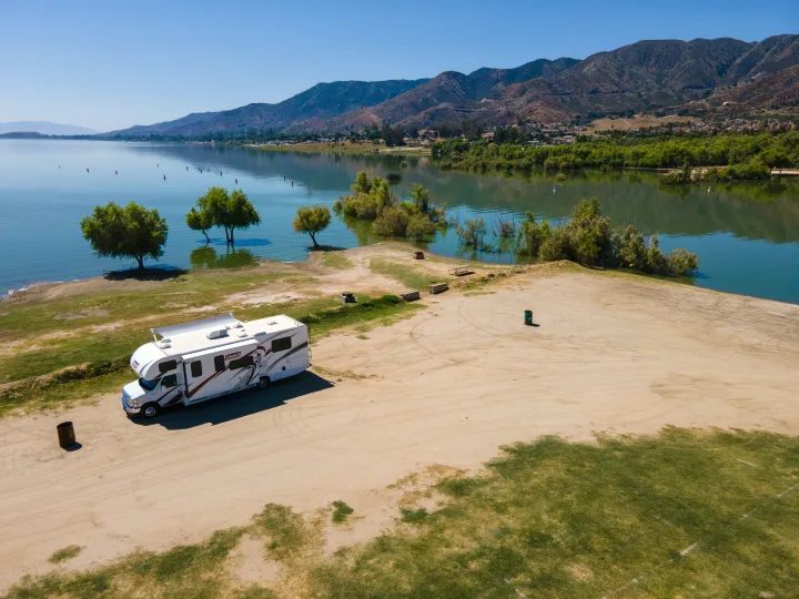Scenic RV rental parked on a sandy shore by a serene lake, featuring trees partly submerged and majestic mountains in the distance.. Thor Coleman 2023