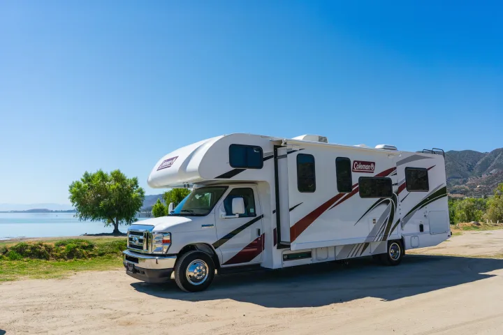 White Coleman Class C RV rental parked at a lakefront with mountains under a clear blue sky.. Thor Coleman 2023