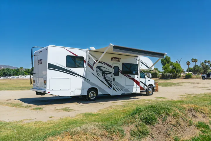 A white Coleman RV rental with its awning extended is parked on a sunny dirt lot, perfect for outdoor travel.. Thor Coleman 2023